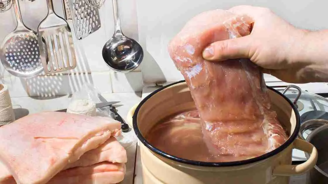 Hand submerging raw pork into a brine solution in a pot on a kitchen counter.
