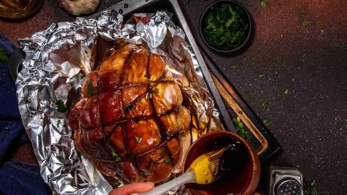 Glazed ham in a roasting pan being basted with a honey glaze, garnished with chopped parsley.