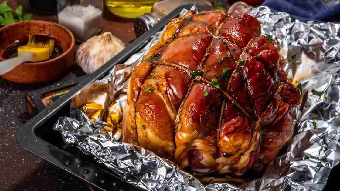 A glazed and smoked ham resting on foil in a roasting pan.