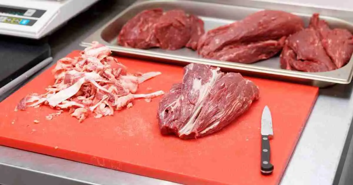 Trimmed-beef Trimmed beef and discarded fat on a red cutting board, with more prepared meat in the background, showcasing the prep work for BBQ or smoking.