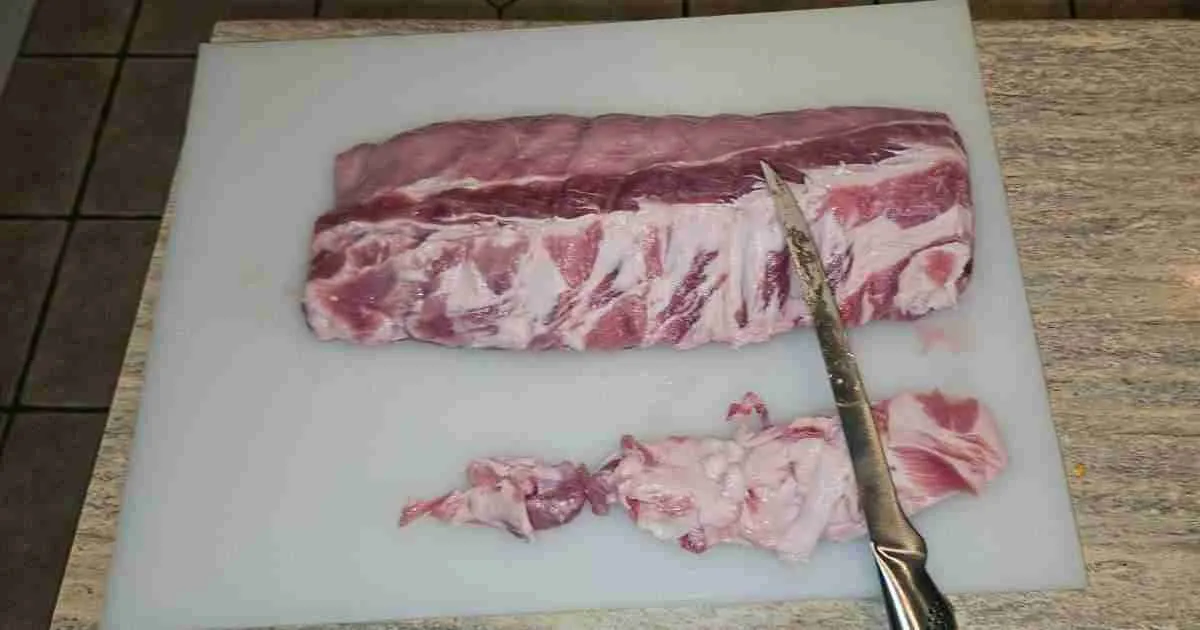 Trimmed-pork-ribs Pre-trimmed rack of pork ribs on a white cutting board with a chef's knife, showing the start of fat removal for even smoking and flavor enhancement.