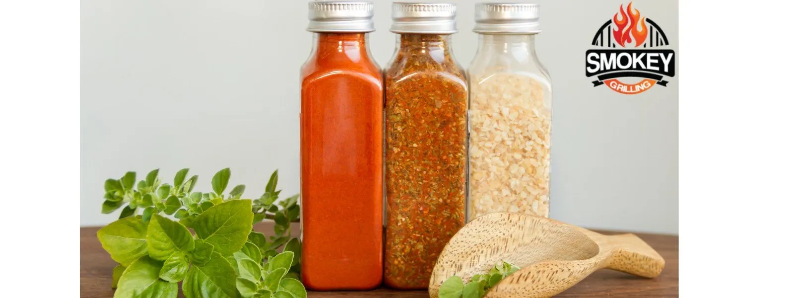 Three glass jars filled with different spices, next to fresh herbs and a wooden spoon, with the Smokey Grilling logo in the top right corner.